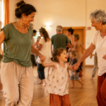 Biodanza en famille : quand la danse réconcilie les générations Famille multigénérationnelle pratiquant la biodanza dans une salle lumineuse, avec un parent, un enfant et une grand-mère dansant pieds nus dans une ambiance chaleureuse et bienveillante.