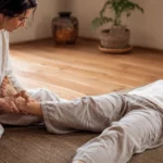 Séance de Wuo-Taï dans un espace de bien-être lumineux : une praticienne assise au sol tient délicatement le pied d’une personne allongée sur un tapis, illustrant cette approche douce inspirée de l’ostéopathie et de la danse.