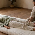 Wuo-Taï à deux : l'art de donner et recevoir dans une danse corporelle Séance de Wuo-Taï à deux sur futon : un praticien transmet un mouvement doux par le contact des jambes dans un espace chaleureux aux sols en bois et à la lumière naturelle.
